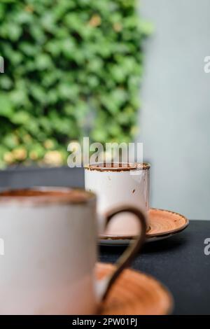 Konzentriere dich auf die andere Seite des Espressos. Zwei Tassen türkischer schwarzer schaumiger Kaffee auf einem Tisch aus schwarzem Stein mit Zweigen von wildem Efeu im Hintergrund, bizet Stockfoto