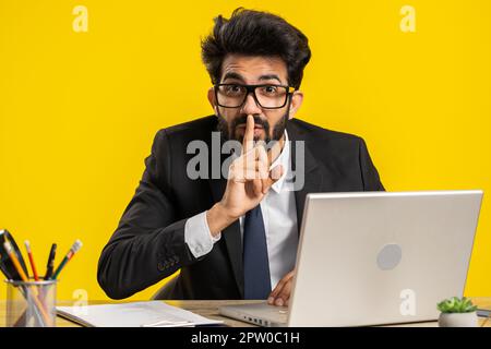 Sei bitte still. Indischer Geschäftsmann drückt Zeigefinger auf Lippen macht Stille Gestenzeichen, die am Arbeitsplatz nichts verraten. Hindu-Mann, der an einem Laptop arbeitet, isoliert auf gelbem Hintergrund Stockfoto