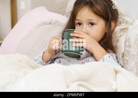 Nettes, süßes, liebes, kleines, dunkelhaariges Vorschulmädchen, trinkt heißen Tee oder Medizin aus grüner Tasse, liegt im Pyjama in einem weichen Bett. Genesung, Gesundheitsfürsorge. Stockfoto