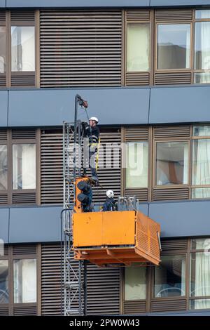 Wartungstechniker auf einer Hängevorrichtung oder Hebebühne, die Reparaturarbeiten an der Fassade eines modernen mehrstöckigen Bürogebäudes und Fensters durchführen Stockfoto