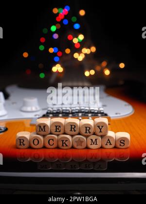 Wooden blocks with the inscription Merry Rock Mas on an old used vintage guitar with colorful Christmas lights bokeh background Stock Photo