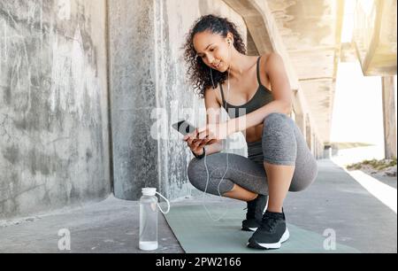 Wie sieht meine Gesundheitsstatistik aus? Aufnahme einer attraktiven jungen Frau in der ganzen Länge, die sich während ihres Workouts im Freien in der Hocke ruht und ihr Handy benutzt Stockfoto