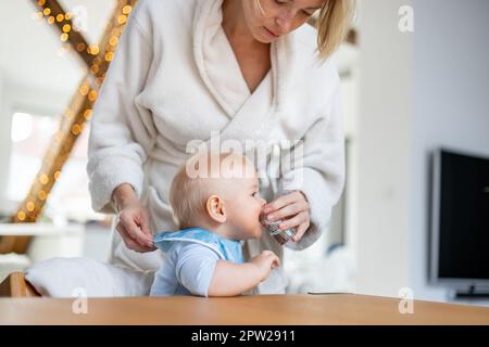 Freizeitkonzept für häusliche Momente. Säuglingsjunge trinkt Wasser aus kleinem Glas mit Hilfe seiner Mutter im kuscheligen Bademantel Stockfoto