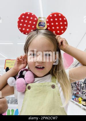 Ein kleines Mädchen im Laden probiert süße rote Minnie-Maus-Ohren mit Punktmuster Stockfoto