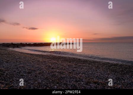 Sonnenaufgang am Kiesstrand mit Wellenbrecher. Unendlicher Hintergrund, orange und rötliche Farben, leerer Strand, kleine Steine. Moncofar Stockfoto