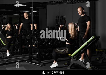 Fitnesstrainer führt persönliches Training für ein Mädchen mit Hantel vor dem Spiegel durch Stockfoto