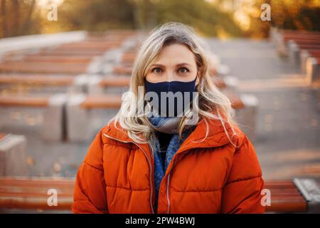 Blonde weibliche in stilvollen Oberbekleidung und Stoff Maske Blick auf Kamera während der Pandemie in der Nähe von Bänken im Park Stockfoto