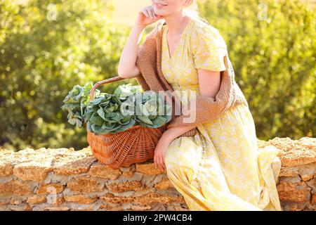 Nicht wiedererkennbare junge Frau, die sich an einem sonnigen Sommertag auf einem Korb mit frischem Kohl und einem berührenden Gesicht lehnt und sich auf einem Ziegelzaun ruht Stockfoto