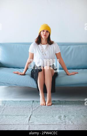 Seitenansicht der barfuß-Frau in trendigen Kleidern Blick auf Kamera auf blauem Sofa vor grauem Hintergrund Stockfoto