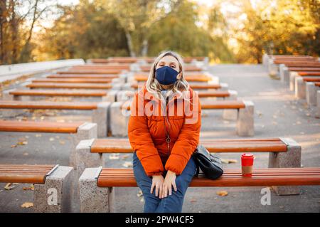 Blonde Frau in stilvoller Oberbekleidung und Stoffmaske, die auf die Kamera schaut, während sie während der Pandemie auf den Bänken im Park sitzt Stockfoto