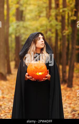 Junge Frau in schwarzem Umhang mit gruseligen Jack o Laterne Und auf den Halloween-Tag im Herbstwald wegschauen Stockfoto