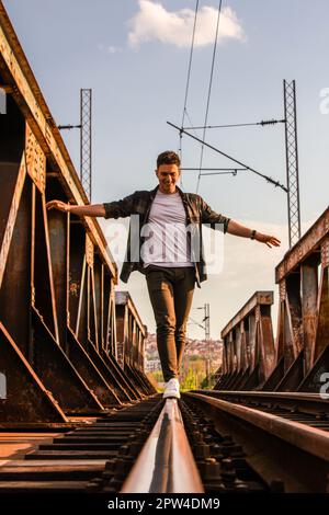 Ein kaukasischer Teenager, der vorsichtig auf Bahngleisen auf einer rostigen Eisenbahnbrücke mit einer verschwommenen Stadt im Hintergrund spaziert Stockfoto