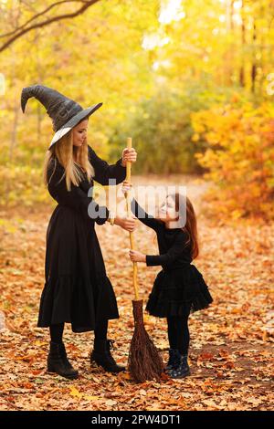 Ganzkörperfrau und Mädchen in Hexenkostümen fegen verwelkte Blätter vom Weg mit Besen während der Halloween-Feier im Herbstwald Stockfoto