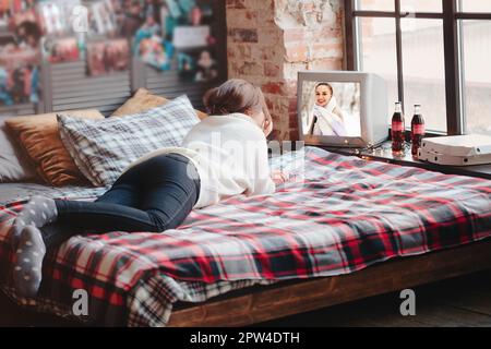 Unkenntliche Frau, die sich auf einem bequemen Bett mit karierter Decke entspannt und zu Hause Retro-TV guckend Stockfoto