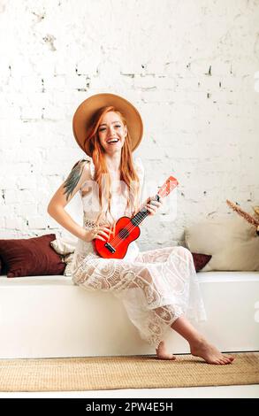 Ganzkörper-glückliche junge Frau in Boho-Kleid und Hut lächelnd und Ukulele spielend, während sie zu Hause auf der Bank gegen die weiße Ziegelwand sitzt Stockfoto