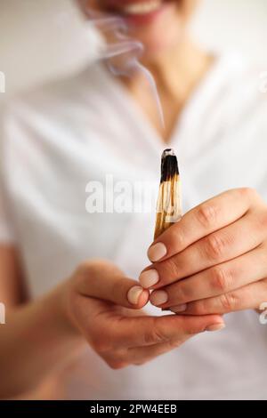 Stimmungsvolles Foto einer lächelnden, weiblichen SPA-Mitarbeiterin in weißer Uniform mit brennendem palo santo Räucherstäbchen und langsam aufsteigendem Rauch. Spirituell Stockfoto