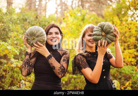 Zwei Freundinnen halten gruselige grüne Kürbisse vor ihrem Gesicht und stehen isoliert vor dem Hintergrund des Herbstwaldes. Herbstferien, halloween Stockfoto