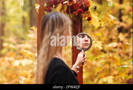 Malerisches Foto eines jungen, wunderschönen Mädchens mit Abendschminke in schwarzem Hexenkostüm mit Spiegel und Blick auf ihn während der halloween-Fotosession Stockfoto