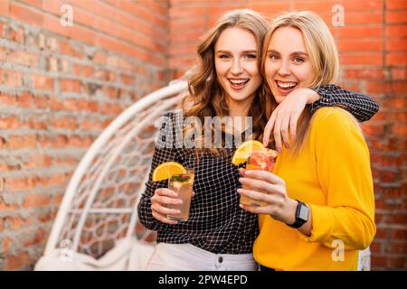 Zwei junge Frauen trinken im Freien gesunde Getränke. Glückliche Freundinnen mit Zitruscocktails auf der Dachparty Stockfoto