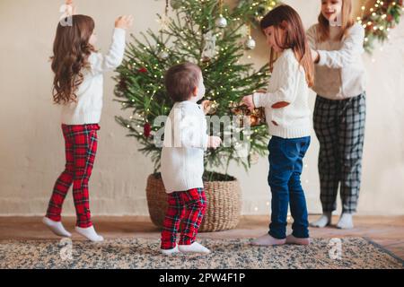 Fröhliche vier kleine Geschwister, drei Schwestern und kleiner Bruder in warmen Strickpullis versammelten sich um Tannenbaum im Wohnzimmer und schmückten es mit Stockfoto