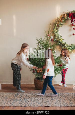 Fröhliche vier kleine Geschwister, drei Schwestern in warmen Strickpullis, die sich im Wohnzimmer um Tannenbaum versammelten und es mit Spielzeug und glänzenden Farben dekorierten Stockfoto