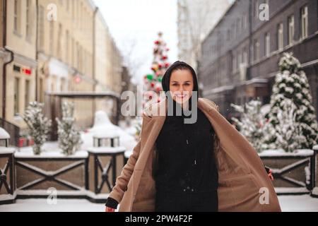 Junge positive Frau mit lustigem Lächeln im Gesicht steht draußen in beigefarbenem Mantel und schwarzem Schal auf dem Kopf und zittert vor kaltem Winterwetter Stockfoto