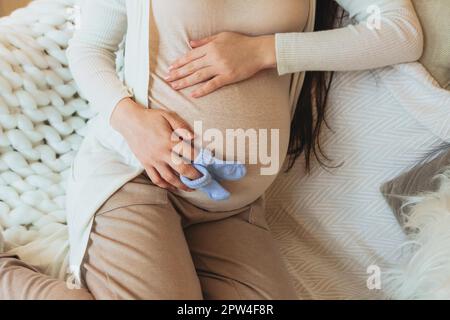 Mutterschafts- und Schwangerschaftskonzept. Verkürztes Foto einer schwangeren Frau, die ein Paar süße blaue Babysocken für das ungeborene Kind auf dem Bauch hält, während sie sich im entspannt Stockfoto