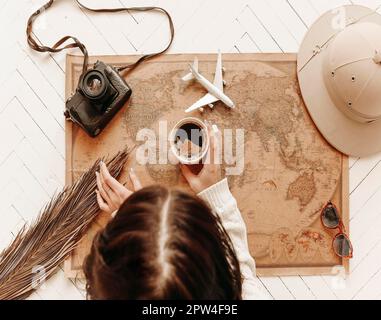 Draufsicht einer Frau, die eine Tasse schwarzen Kaffee auf einer Weltkarte im Vintage-Look hält, mit Reisezubehör, Retro-Kamera, Spielzeugflugzeug, das auf der Karte liegt Stockfoto