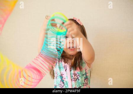 Das niedliche, niedliche kleine Mädchen spielt mit einer magischen Quelle aus Plastik, hält elastisches Spielzeug und lächelt vor der Kamera. Glückliches Kind isoliert auf Beige Stockfoto