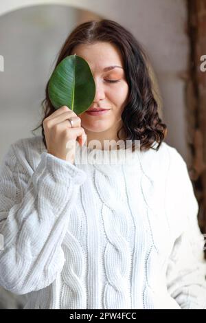 Porträt eines jungen braunen Mädchens mit wunderschön gestyltem Haar in weißem, warmgestricktem Pullover, das ihr Auge ruhig mit grünem Pflanzenblatt bedeckt Stockfoto