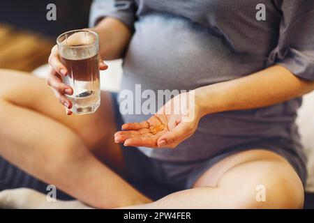 Nahrungsergänzungsmittel während der Schwangerschaft. Junge, glückliche, schwangere Frau, die pränatale Vitamine einnimmt, ein Glas Wasser und eine Pille hält, während sie zu Hause im Bett sitzt. Stockfoto