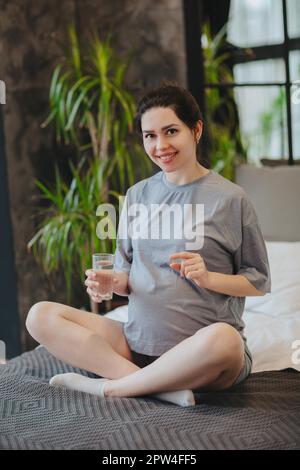 Nahrungsergänzungsmittel während der Schwangerschaft. Junge, glückliche, schwangere Frau, die pränatale Vitamine einnimmt, ein Glas Wasser und eine Pille hält, während sie zu Hause im Bett sitzt. Stockfoto