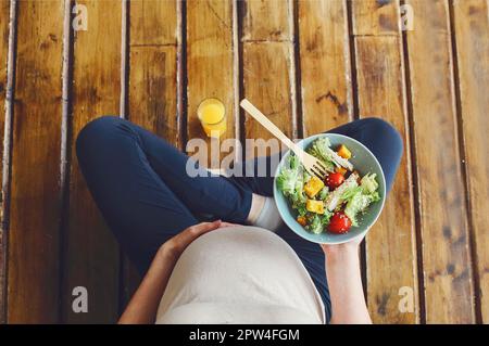 Blick von oben auf schwangere Frau mit frischer Salatschüssel und Orangensaft, werdende Mutter hat gesundes Mittagessen, während sie auf Holzboden sitzt Stockfoto