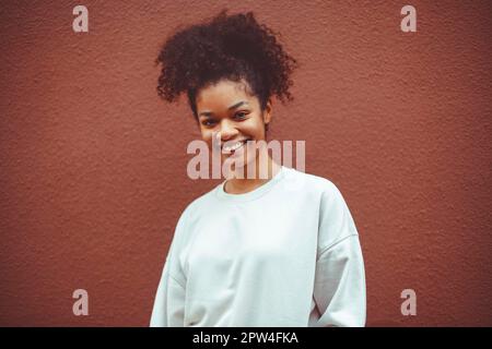 Reines Glück. Süßes, glückliches afroamerikanisches Mädchen mit lockiger Frisur, das einen weißen Pullover trägt und gegen die braune Wand posiert. Lächelnde schwarze junge Frau Stockfoto