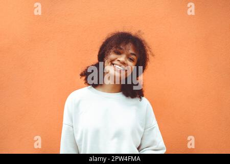 Reines Glück. Süßes, glückliches afroamerikanisches Mädchen mit lockiger Frisur in einem weißen Pullover, das an der orangefarbenen Wand posiert. Lächelnde schwarze junge Frau Stockfoto