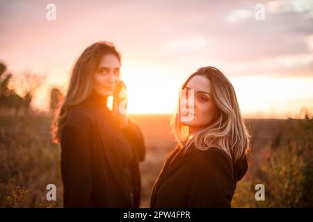 Zwei fröhliche Frauen, die Händchen halten, gehen bei Sonnenuntergang durch das Herbstfeld und lächeln, sorgenfreie weibliche beste Freunde, die den Abend genießen Stockfoto