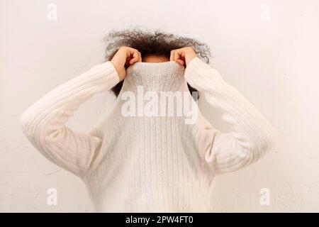 Afrikanisch geschwungenes weibliches Abdeckgesicht mit warmweißem Pullover isoliert auf hellem Studiohintergrund, weiblich mit Polokragen-Pullover, kann nicht Stockfoto