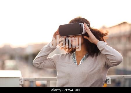 Technologie der Zukunft. Junge, moderne Mixed-Race-Frau in VR-Brille Headset erkunden virtuelle Realität im Freien mit Stadtbild auf dem Hintergrund, weibliche versuchen Stockfoto