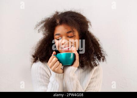 Fröhliches, entzückendes, lockiges, afroamerikanisches Mädchen mit geschlossenen Augen, das eine Tasse Tee genießt und sich in der kalten Wintersaison entspannt und warm gestrickt trägt Stockfoto