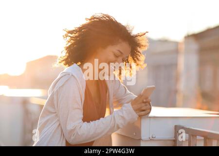 Außenporträt einer fröhlichen afroamerikanischen Frau mit Kopfhörern, die ihr Smartphone benutzt und am Morgen auf dem Dach fröhlich lächelt Stockfoto