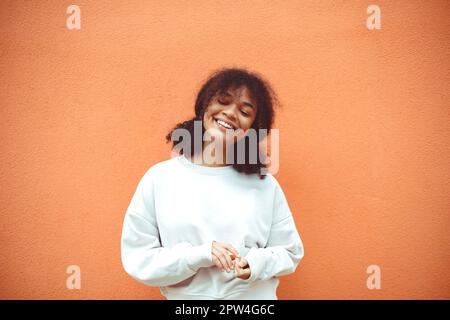 Reines Glück. Süßes, glückliches afroamerikanisches Mädchen mit lockiger Frisur, das einen weißen Pullover trägt und mit geschlossenen Augen an der orangefarbenen Wand posiert. Lächelnd Stockfoto