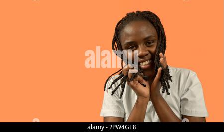 Nahaufnahme des Porträts einer glücklichen jungen Afrikanerin, die mit einem großen Lächeln in die Kamera schaut, mit ihren Haaren mit den Händen spielt, in einem weißen T-Shirt und einem Brötchen Stockfoto