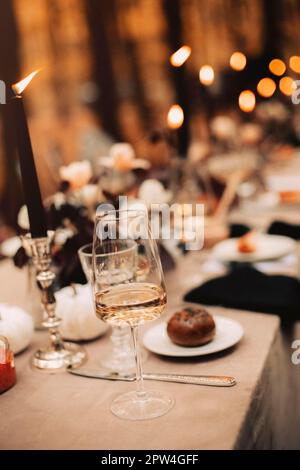 Selektiver Fokus auf hohes Glas mit Weißwein im Vordergrund, vor unscharfem Hintergrund eines ungereinigten Esstisches nach festlicher Sitzbank Stockfoto