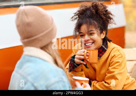 Zwei glückliche, multirassische Freundinnen sitzen draußen, trinken heißen Tee und unterhalten sich am Herbsttag beim Treffen im Park, lächeln weibliche beste Freundinnen Stockfoto