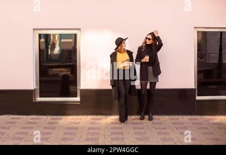Zwei stilvolle weibliche Freundinnen in trendigen Kleidern, die beim Spaziergang durch die Stadt am Wochenende nette Gespräche führen und Kaffee zum Mitnehmen trinken. Stockfoto