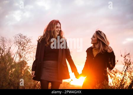 Zwei fröhliche Frauen, die Händchen halten, gehen bei Sonnenuntergang durch das Herbstfeld und lächeln, sorgenfreie weibliche beste Freunde, die den Abend genießen Stockfoto