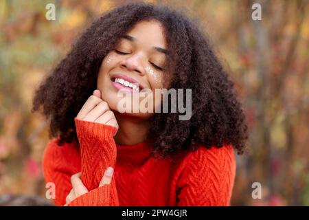 Junge, glückliche, überfreuliche afroamerikanische Frau mit lockigem Haar, die die Augen geschlossen hält, lachend und Spaß dabei hat, im Herbstwald zu stehen und zu genießen Stockfoto
