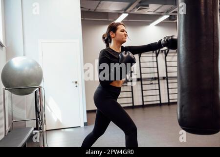 Eine starke latino-Boxerin springt in einen riesigen Boxsack im Fitness-Studio. Sportliches Mädchen trainiert Muay Thai Boxen für Bodybuilding und gesund Stockfoto