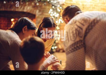 Glückliche Eltern mit Kindern, die die Weihnachtszeit zu Hause genießen. Vater, Mutter und Kinder in der Nähe des Kamins mit Weihnachtsbeleuchtung und Familientoast Stockfoto