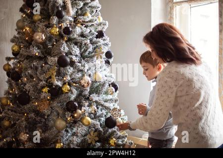 Kleiner Vorschulkind, der Großmutter hilft, zu Hause Weihnachtsbaum zu schmücken, Großmutter und kleiner Enkel, die zusammen Weihnachtsbaum schmücken, Junge Stockfoto